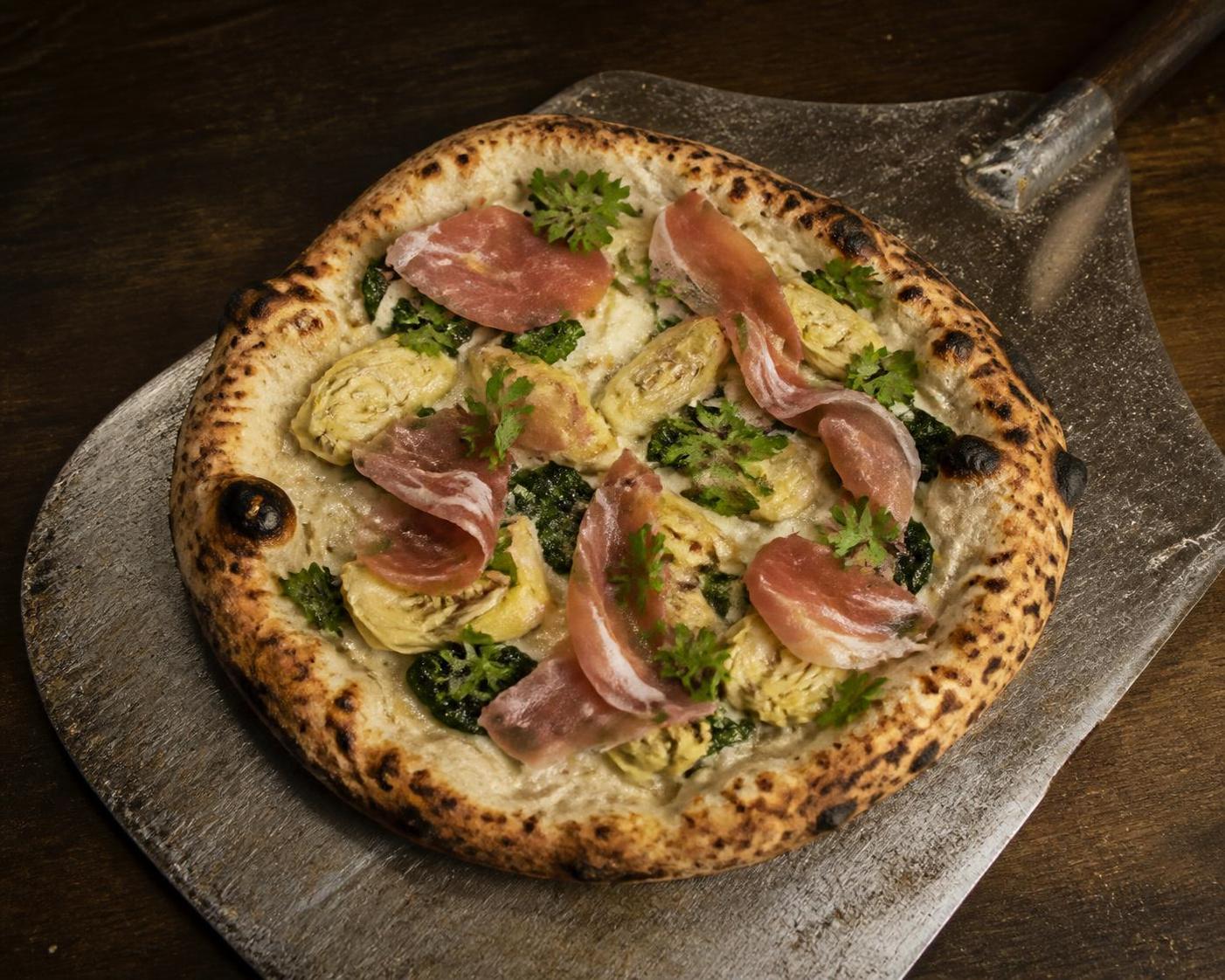Wood-fired pizza with artichoke, greens, and prosciutto on a peel